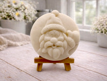 Load image into Gallery viewer, A circular handmade vegan soap featuring a detailed high-relief carving of a jolly Father Christmas waving, with a textured beard and a classic Santa hat, displayed on a miniature wooden easel.
