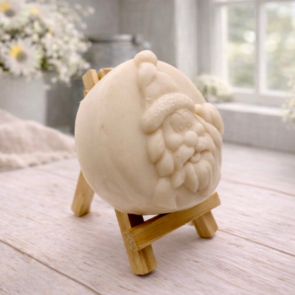 A circular handmade vegan soap featuring a detailed high-relief carving of a jolly Father Christmas waving, with a textured beard and a classic Santa hat, displayed on a miniature wooden easel.