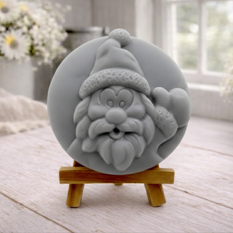 A circular handmade vegan soap featuring a detailed high-relief carving of a jolly Father Christmas waving, with a textured beard and a classic Santa hat, displayed on a miniature wooden easel.