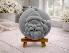 Load image into Gallery viewer, A circular handmade vegan soap featuring a detailed high-relief carving of a jolly Father Christmas waving, with a textured beard and a classic Santa hat, displayed on a miniature wooden easel.
