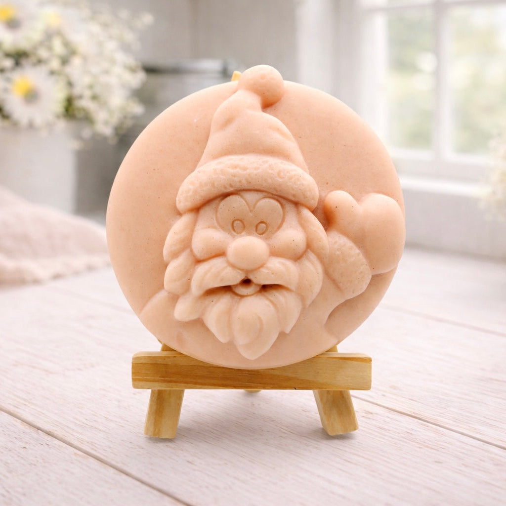 A circular handmade vegan soap featuring a detailed high-relief carving of a jolly Father Christmas waving, with a textured beard and a classic Santa hat, displayed on a miniature wooden easel.