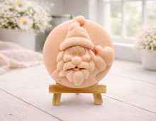 Load image into Gallery viewer, A circular handmade vegan soap featuring a detailed high-relief carving of a jolly Father Christmas waving, with a textured beard and a classic Santa hat, displayed on a miniature wooden easel.
