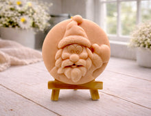Load image into Gallery viewer, A circular handmade vegan soap featuring a detailed high-relief carving of a jolly Father Christmas waving, with a textured beard and a classic Santa hat, displayed on a miniature wooden easel.

