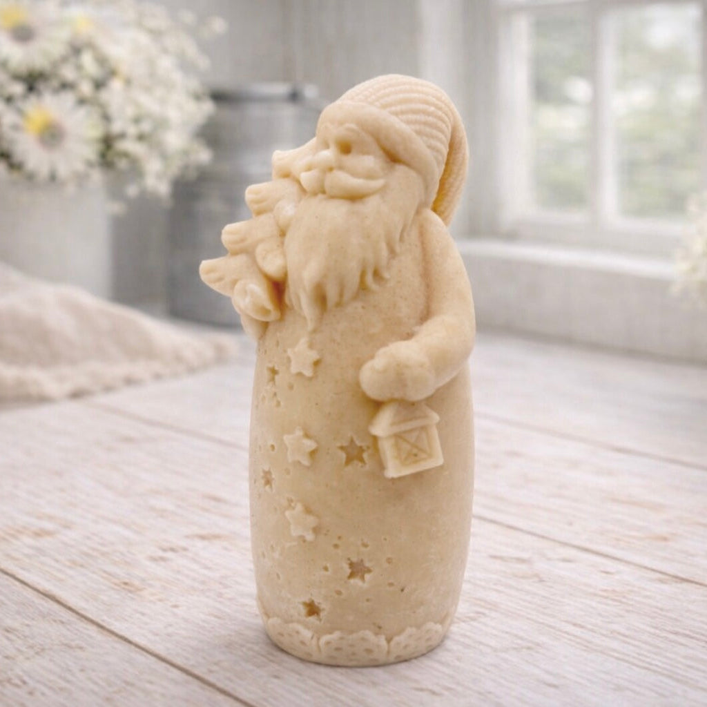 handmade vegan soap sculpted into a tall figure of Father Christmas wearing star-patterned robes, holding a small Christmas tree in one arm and a lantern in the other, set in a bright farmhouse window.