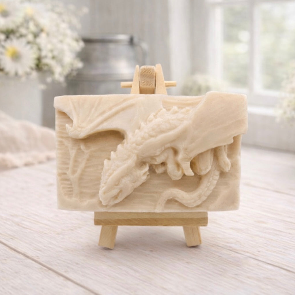 handmade soap bar featuring a detailed 3D relief carving of a dragon, displayed on a small wooden easel in a bright, rustic room with white flowers in the background.