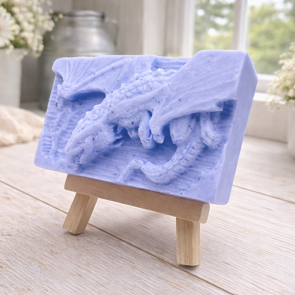 handmade soap bar featuring a detailed 3D relief carving of a dragon, displayed on a small wooden easel in a bright, rustic room with white flowers in the background.