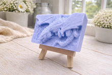 Load image into Gallery viewer, handmade soap bar featuring a detailed 3D relief carving of a dragon, displayed on a small wooden easel in a bright, rustic room with white flowers in the background.

