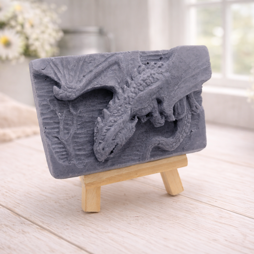 handmade soap bar featuring a detailed 3D relief carving of a dragon, displayed on a small wooden easel in a bright, rustic room with white flowers in the background.