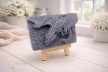 Load image into Gallery viewer, handmade soap bar featuring a detailed 3D relief carving of a dragon, displayed on a small wooden easel in a bright, rustic room with white flowers in the background.
