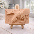 handmade soap bar featuring a detailed 3D relief carving of a dragon, displayed on a small wooden easel in a bright, rustic room with white flowers in the background.