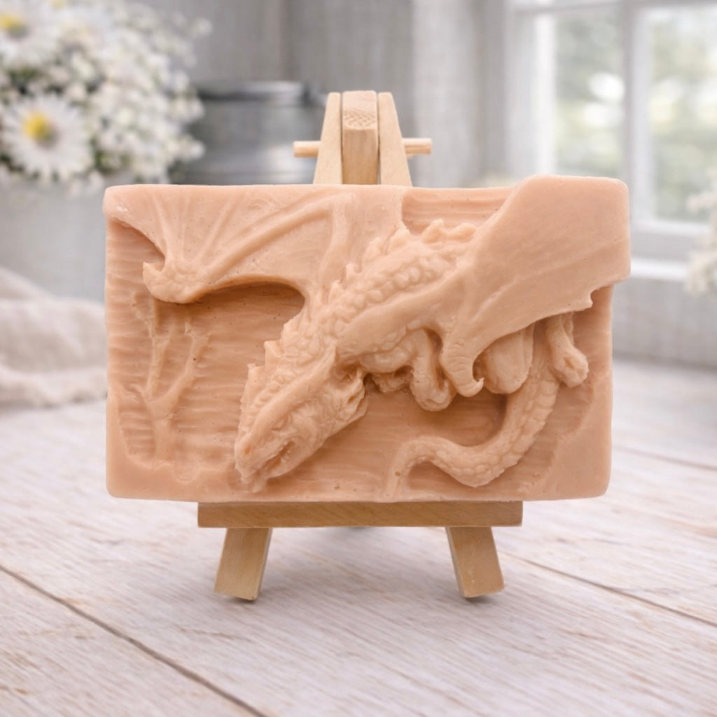 handmade soap bar featuring a detailed 3D relief carving of a dragon, displayed on a small wooden easel in a bright, rustic room with white flowers in the background.
