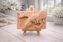 Load image into Gallery viewer, handmade soap bar featuring a detailed 3D relief carving of a dragon, displayed on a small wooden easel in a bright, rustic room with white flowers in the background.
