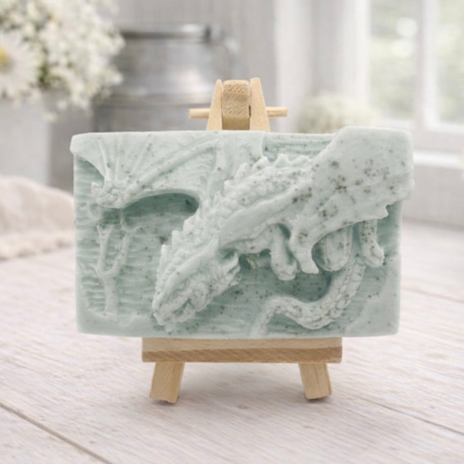 handmade soap bar featuring a detailed 3D relief carving of a dragon, displayed on a small wooden easel in a bright, rustic room with white flowers in the background.