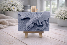 Load image into Gallery viewer, handmade soap bar featuring a detailed 3D relief carving of a dragon, displayed on a small wooden easel in a bright, rustic room with white flowers in the background.
