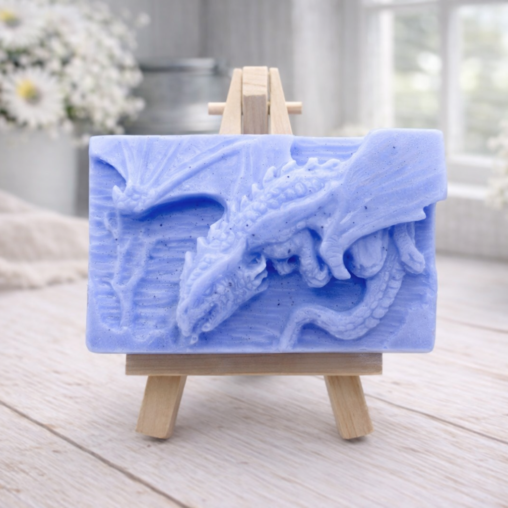 handmade soap bar featuring a detailed 3D relief carving of a dragon, displayed on a small wooden easel in a bright, rustic room with white flowers in the background.