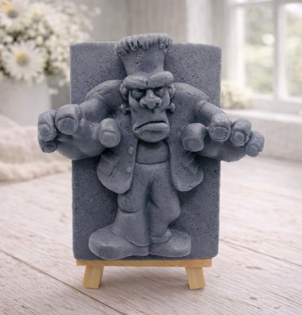 A rectangular handmade vegan soap featuring a high-relief carving of Frankenstein's monster in a classic pose with arms outstretched and a grumpy expression, displayed on a miniature wooden easel.