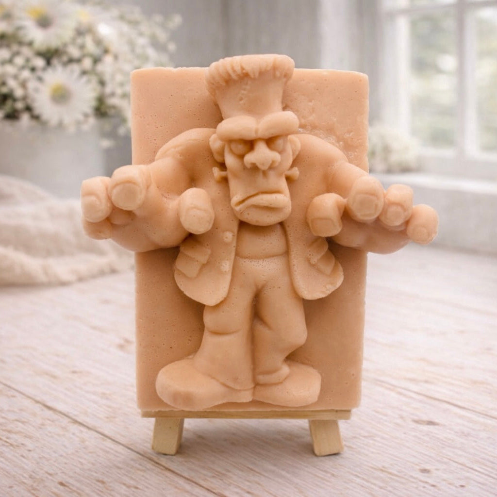 A rectangular handmade vegan soap featuring a high-relief carving of Frankenstein's monster in a classic pose with arms outstretched and a grumpy expression, displayed on a miniature wooden easel.