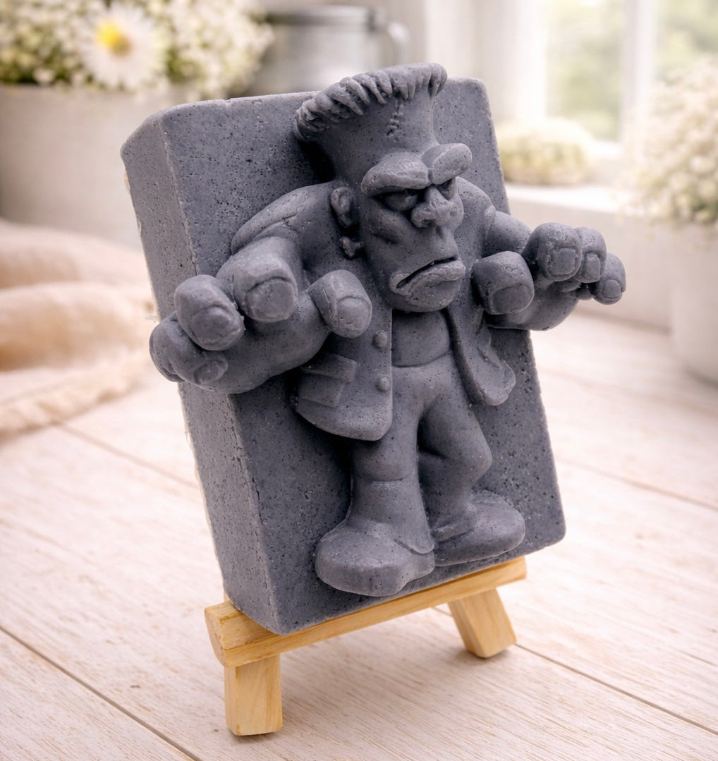 A rectangular handmade vegan soap featuring a high-relief carving of Frankenstein's monster in a classic pose with arms outstretched and a grumpy expression, displayed on a miniature wooden easel.