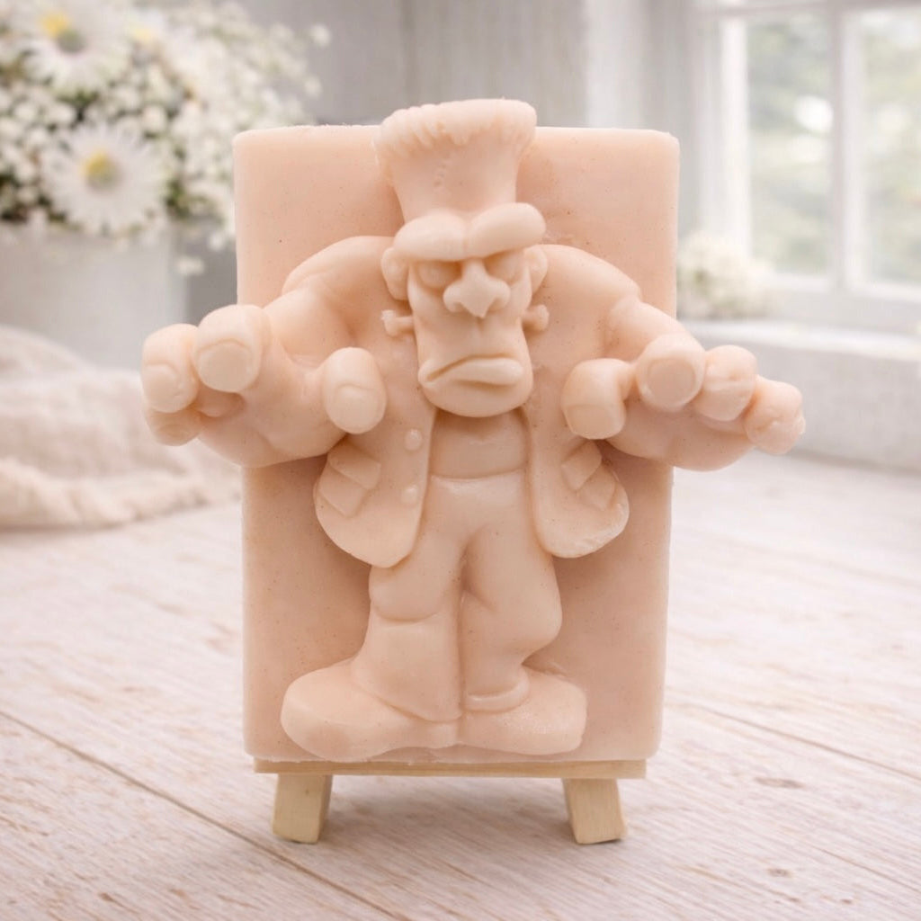A rectangular handmade vegan soap featuring a high-relief carving of Frankenstein's monster in a classic pose with arms outstretched and a grumpy expression, displayed on a miniature wooden easel.