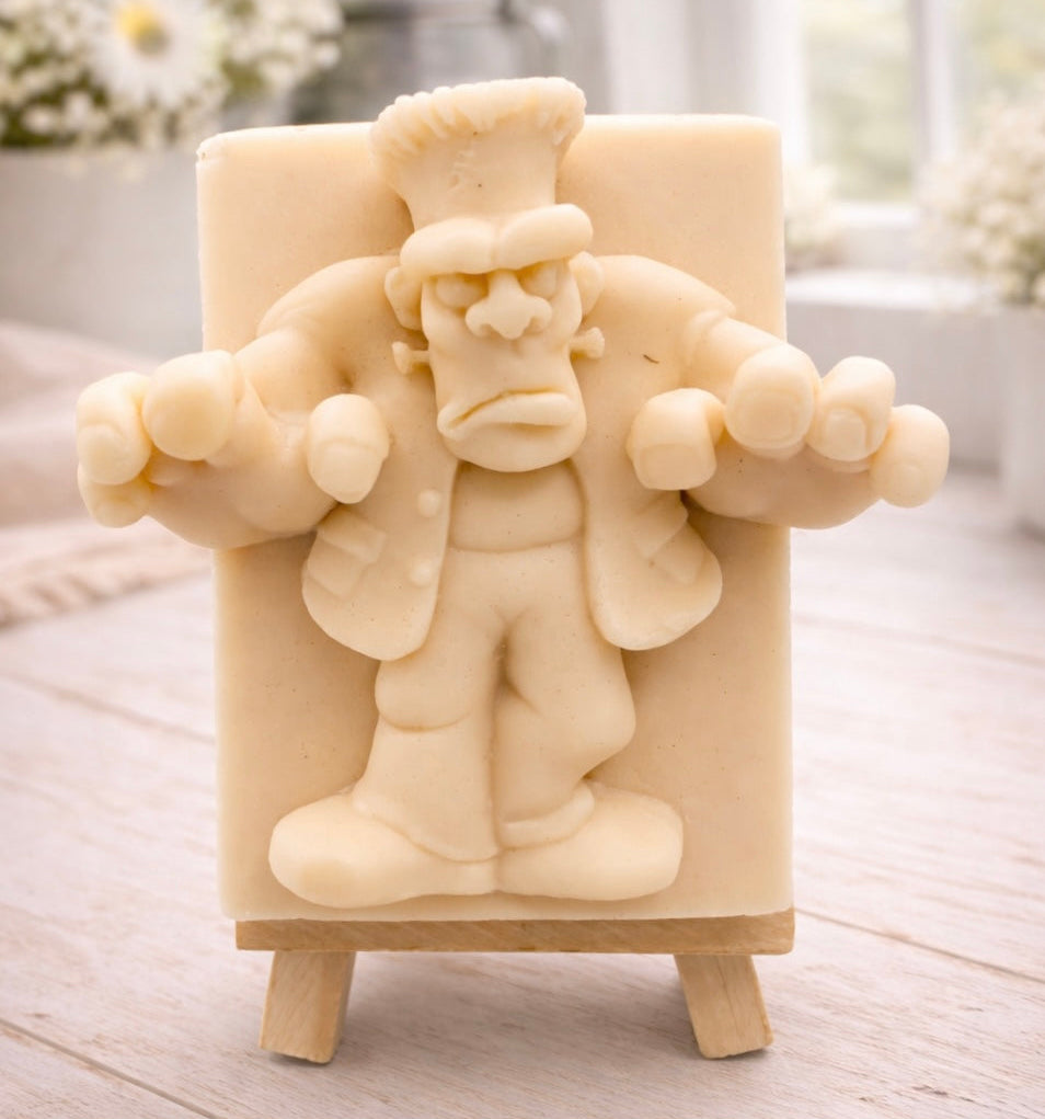 A rectangular handmade vegan soap featuring a high-relief carving of Frankenstein's monster in a classic pose with arms outstretched and a grumpy expression, displayed on a miniature wooden easel.