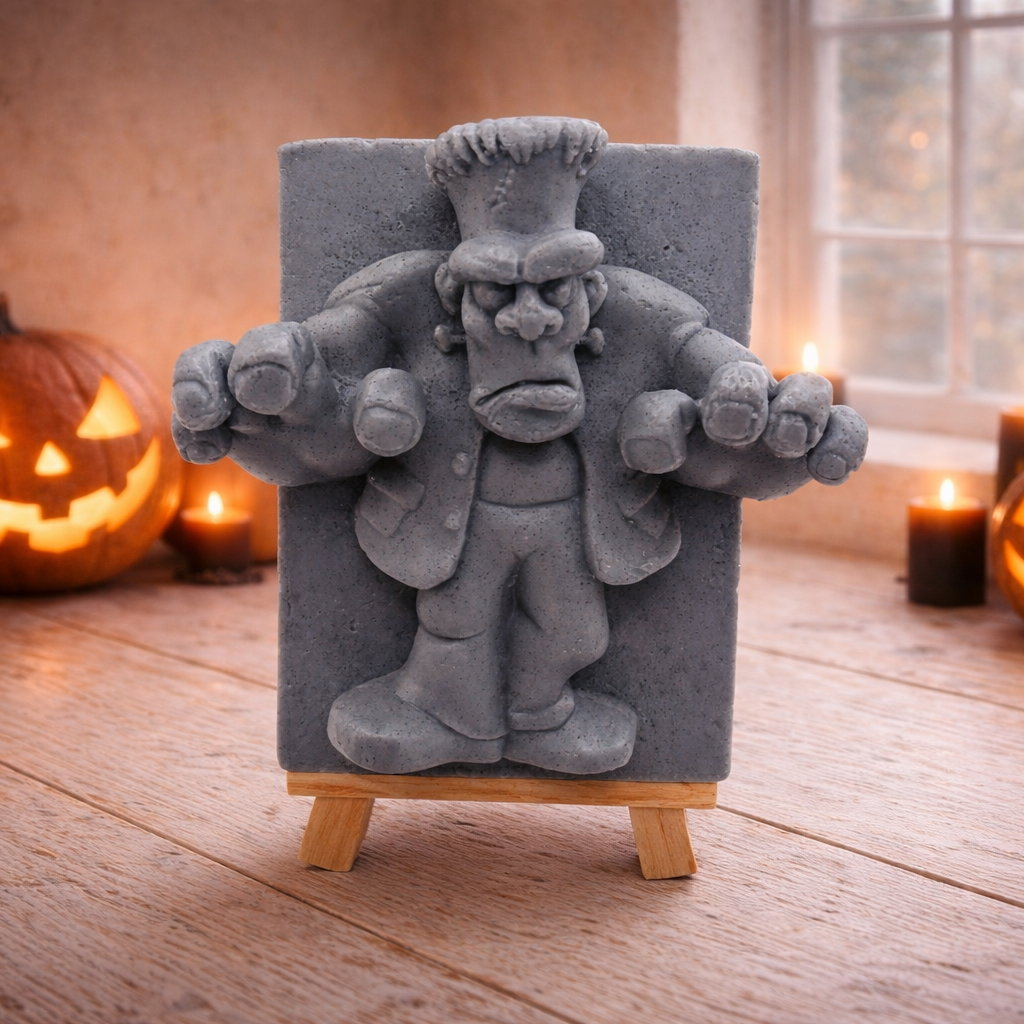 A rectangular handmade vegan soap featuring a high-relief carving of Frankenstein's monster in a classic pose with arms outstretched and a grumpy expression, displayed on a miniature wooden easel.
