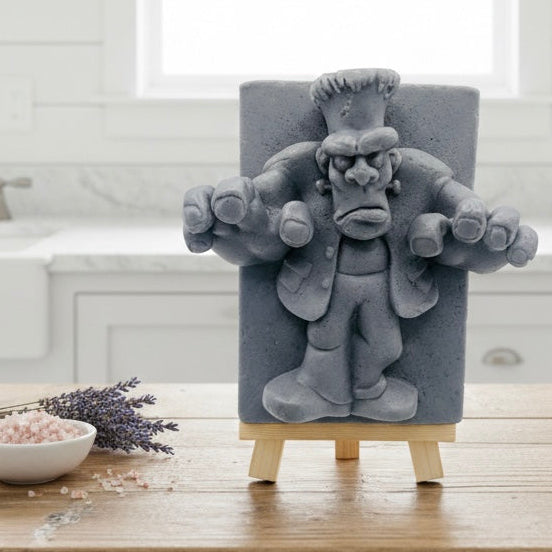 A detailed handmade vegan soap featuring a 3D relief of Frankenstein's monster, displayed on a wooden easel against a whimsical farmhouse decor background.