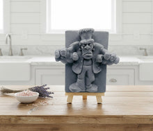 Load image into Gallery viewer, A detailed handmade vegan soap featuring a 3D relief of Frankenstein's monster, displayed on a wooden easel against a whimsical farmhouse decor background.
