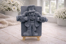 Load image into Gallery viewer, A rectangular handmade vegan soap featuring a high-relief carving of Frankenstein's monster in a classic pose with arms outstretched and a grumpy expression, displayed on a miniature wooden easel.
