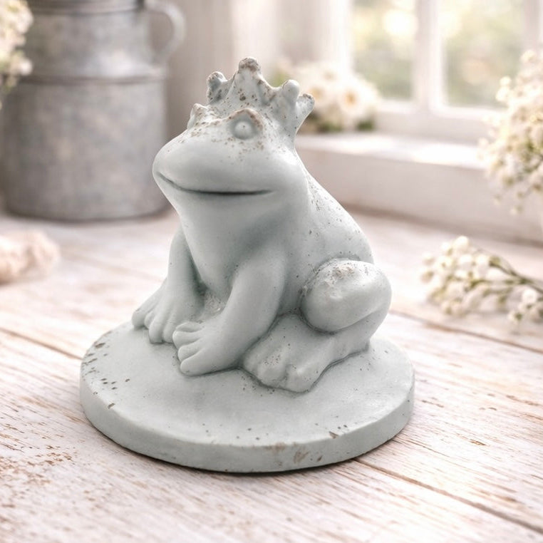 A 3D handmade vegan soap in a pale mint-green tone, sculpted into a sitting frog wearing a crown on its head, positioned on a circular base and surrounded by delicate white flowers in a bright farmhouse setting.