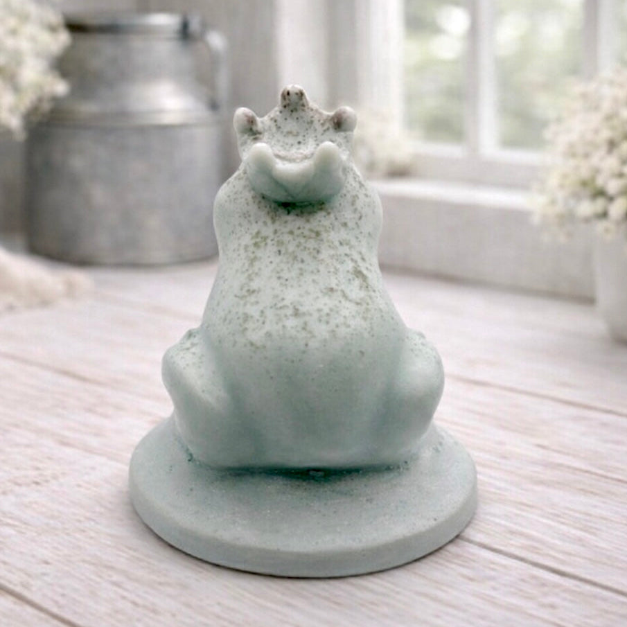A 3D handmade vegan soap in a pale mint-green tone, sculpted into a sitting frog wearing a crown on its head, positioned on a circular base and surrounded by delicate white flowers in a bright farmhouse setting.
