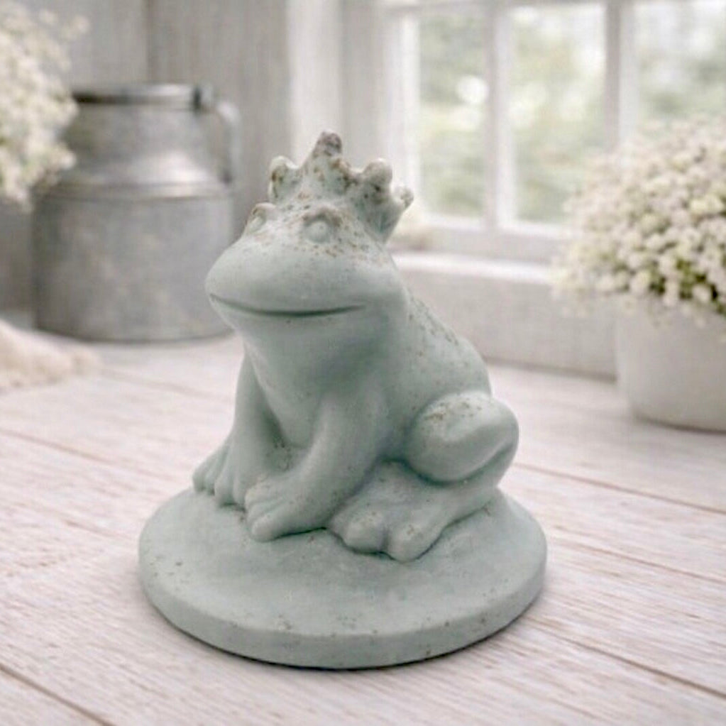 A 3D handmade vegan soap in a pale mint-green tone, sculpted into a sitting frog wearing a crown on its head, positioned on a circular base and surrounded by delicate white flowers in a bright farmhouse setting.