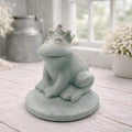 A 3D handmade vegan soap in a pale mint-green tone, sculpted into a sitting frog wearing a crown on its head, positioned on a circular base and surrounded by delicate white flowers in a bright farmhouse setting.