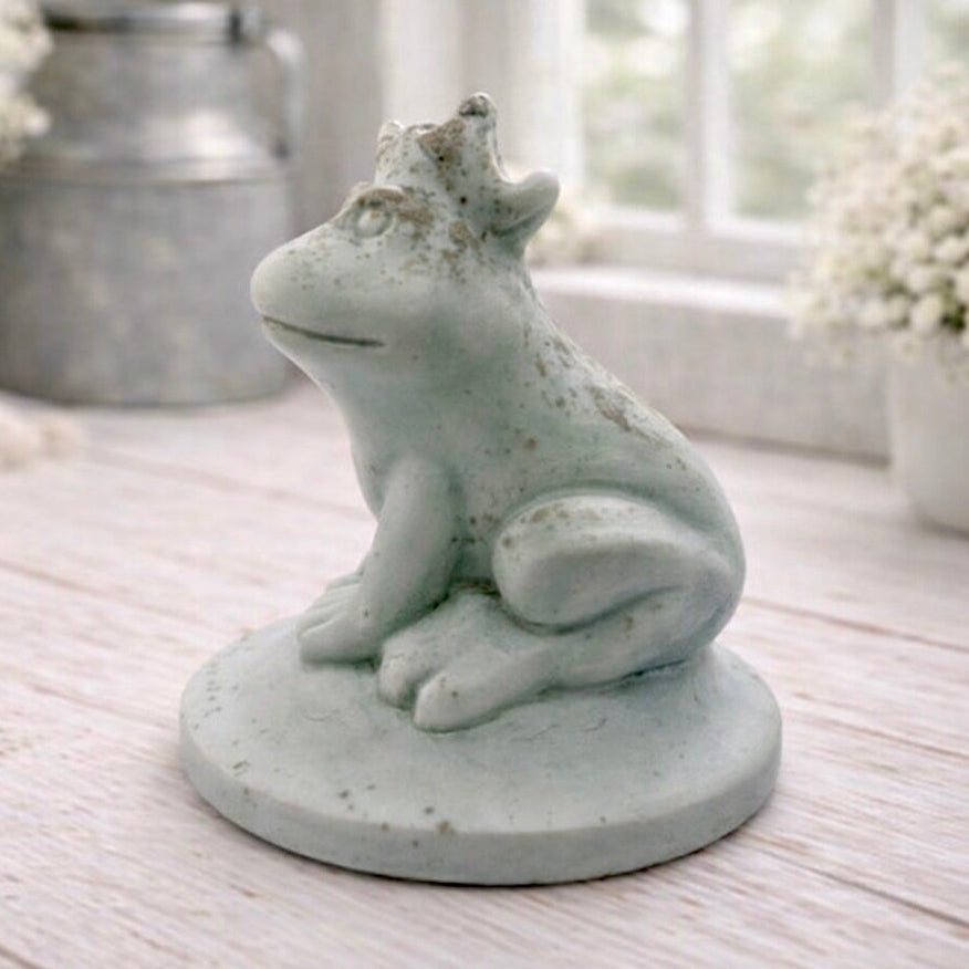 A 3D handmade vegan soap in a pale mint-green tone, sculpted into a sitting frog wearing a crown on its head, positioned on a circular base and surrounded by delicate white flowers in a bright farmhouse setting.