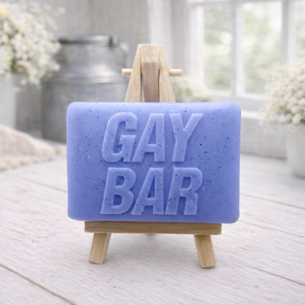 A rectangular handmade vegan soap  featuring the words "GAY BAR" in large, bold, 3D block lettering, displayed on a miniature wooden easel.