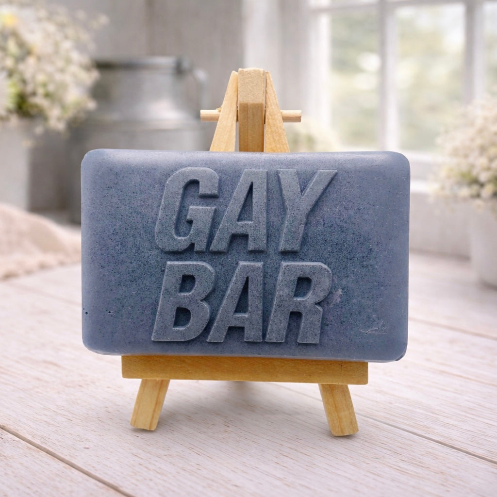A rectangular handmade vegan soap  featuring the words "GAY BAR" in large, bold, 3D block lettering, displayed on a miniature wooden easel.