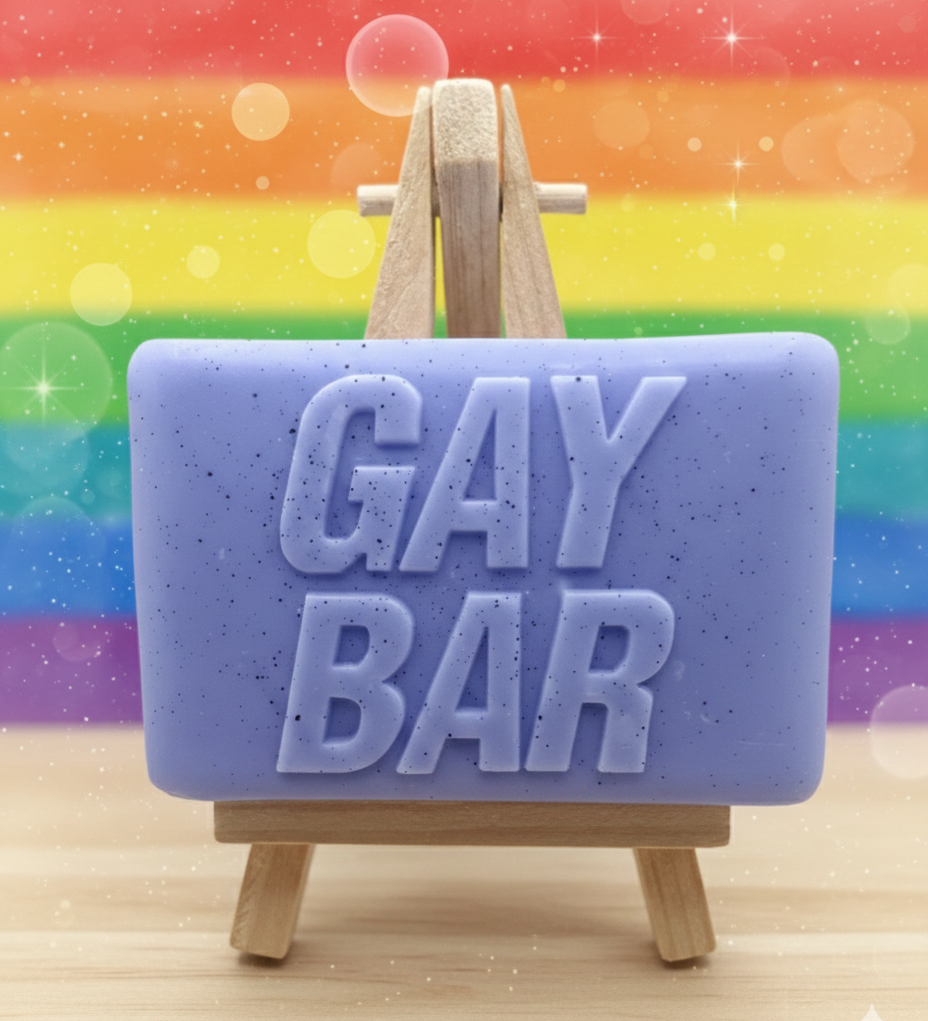 A soap bar with 'GAY BAR' text on a wooden stand against a rainbow background