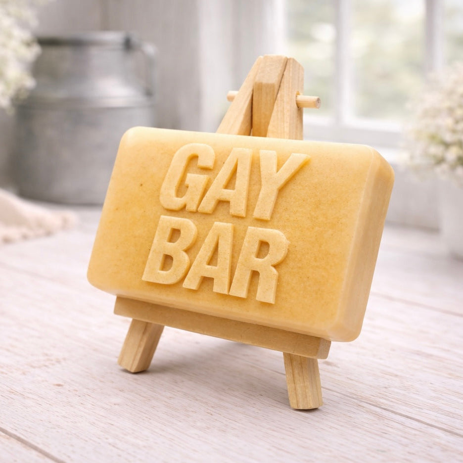 A rectangular handmade vegan soap featuring the words "GAY BAR" in large, bold, 3D block lettering, displayed on a miniature wooden easel.