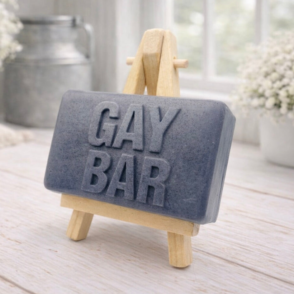 Gray soap bar with 'GAY BAR' text on a wooden stand, placed on a light wooden surface.