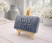 Load image into Gallery viewer, Gray soap bar with 'GAY BAR' text on a wooden stand, placed on a light wooden surface.
