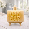 A rectangular handmade vegan soap  featuring the words "GAY BAR" in large, bold, 3D block lettering, displayed on a miniature wooden easel.