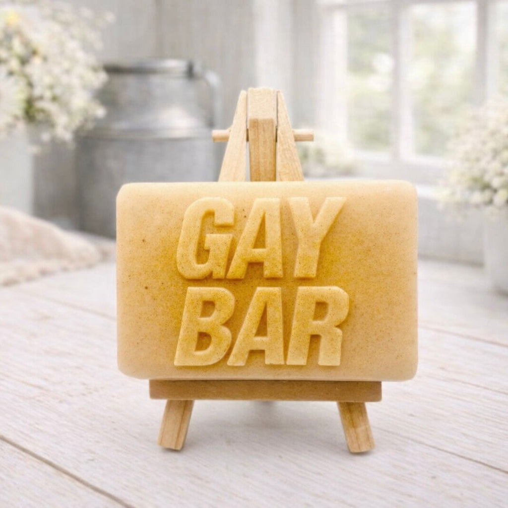 A rectangular handmade vegan soap  featuring the words "GAY BAR" in large, bold, 3D block lettering, displayed on a miniature wooden easel.