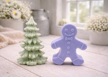 Load image into Gallery viewer, A handmade vegan soap featuring a classic gingerbread man with a smiling face, a bow tie, and decorative icing details on his arms and legs, and a Christmas tree - displayed on a miniature wooden easel.
