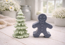 Load image into Gallery viewer, A handmade vegan soap featuring a classic gingerbread man with a smiling face, a bow tie, and decorative icing details on his arms and legs, and a Christmas tree - displayed on a miniature wooden easel.
