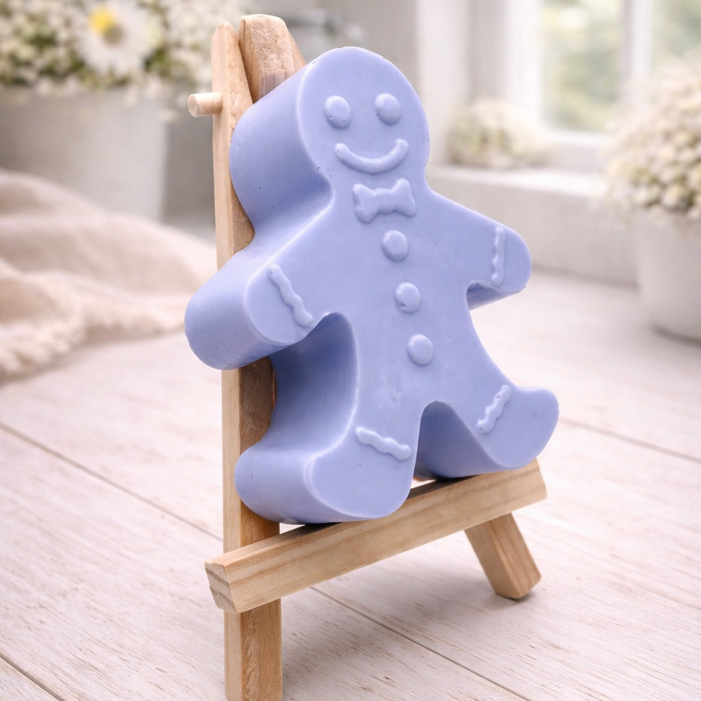 A handmade vegan featuring a classic gingerbread man with a smiling face, a bow tie, and decorative icing details on his arms and legs, displayed on a miniature wooden easel.