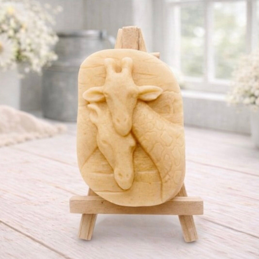 A rectangular handmade vegan soap in a natural tone, featuring a detailed relief of a mother and baby giraffe with their necks intertwined, displayed on a miniature wooden easel against a white wood-panelled background.