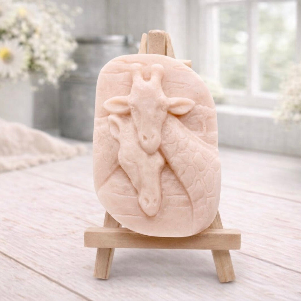 A rectangular handmade vegan soap in a natural tone, featuring a detailed relief of a mother and baby giraffe with their necks intertwined, displayed on a miniature wooden easel against a white wood-panelled background.