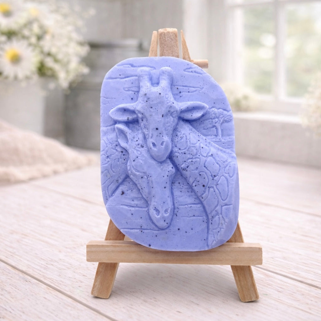 A rectangular handmade vegan soap in a natural tone, featuring a detailed relief of a mother and baby giraffe with their necks intertwined, displayed on a miniature wooden easel against a white wood-panelled background.