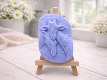 Load image into Gallery viewer, A rectangular handmade vegan soap in a natural tone, featuring a detailed relief of a mother and baby giraffe with their necks intertwined, displayed on a miniature wooden easel against a white wood-panelled background.
