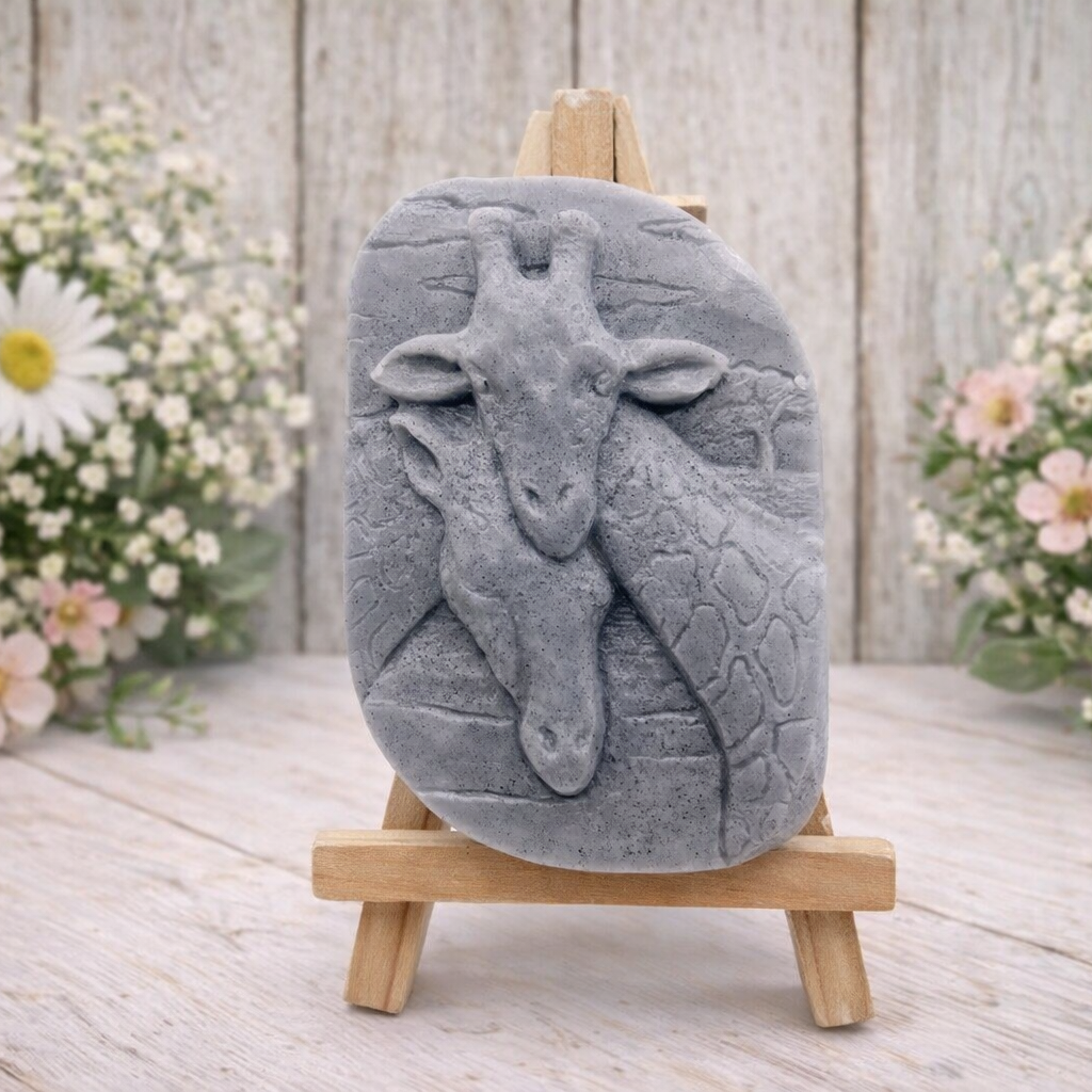 A rectangular handmade vegan soap in a natural tone, featuring a detailed relief of a mother and baby giraffe with their necks intertwined, displayed on a miniature wooden easel against a white wood-panelled background.