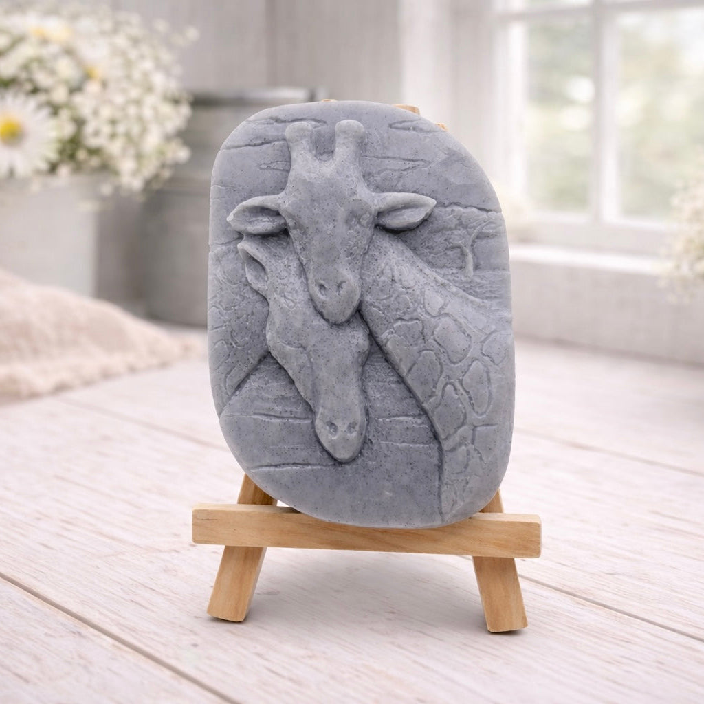 A rectangular handmade vegan soap in a natural tone, featuring a detailed relief of a mother and baby giraffe with their necks intertwined, displayed on a miniature wooden easel against a white wood-panelled background.